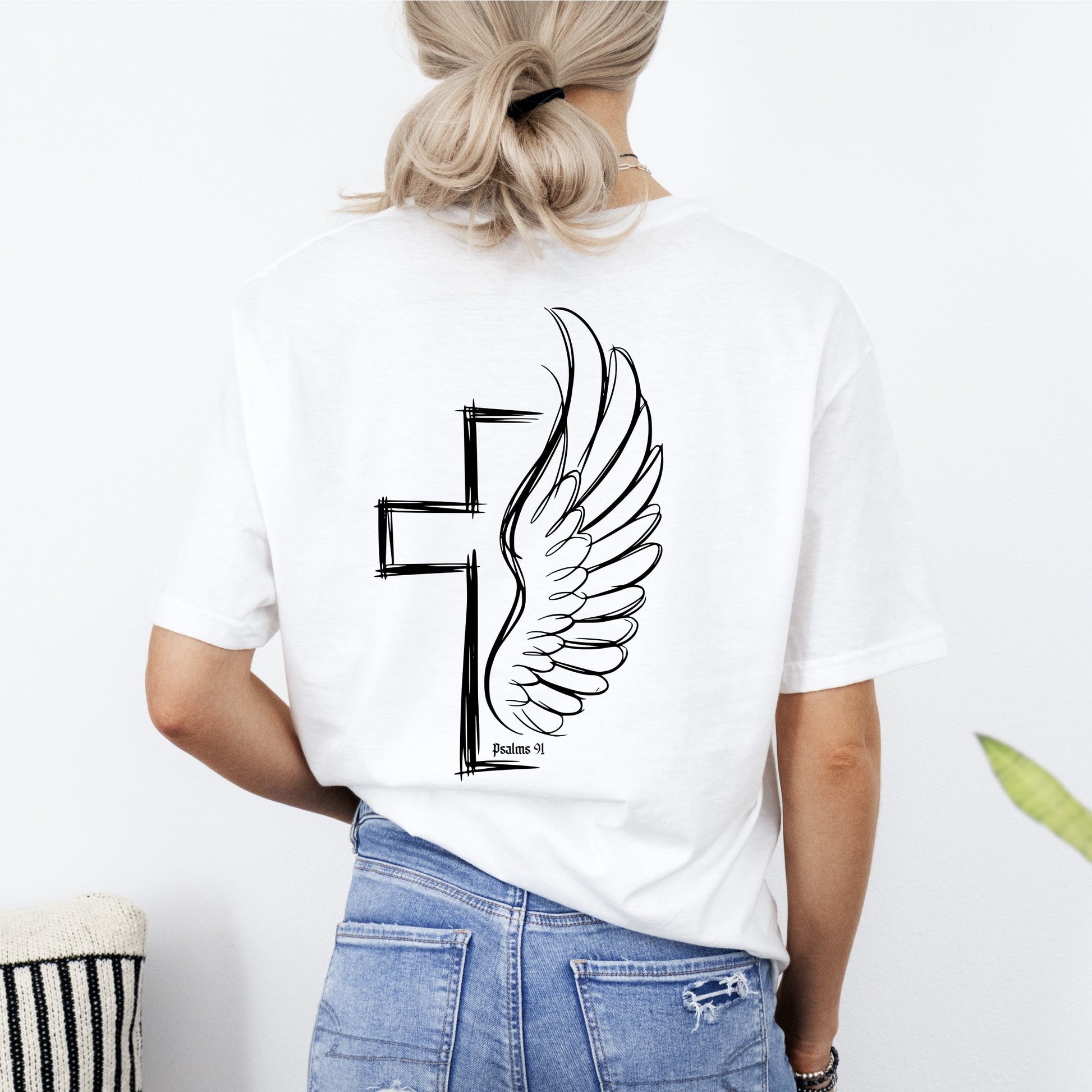 Person wearing a white t-shirt with a black cross and wing design, standing against a plain background.