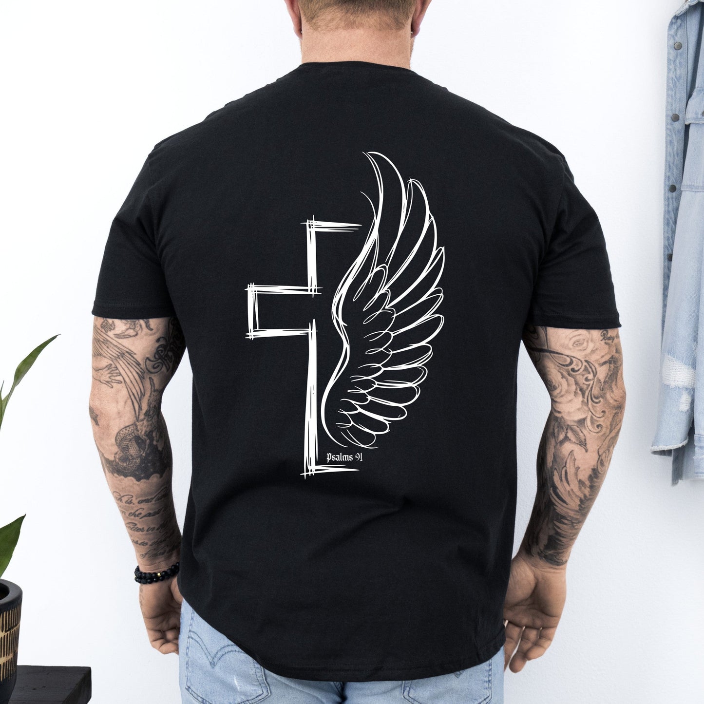 Person wearing a black t-shirt with a winged cross design on the back, standing against a white wall.