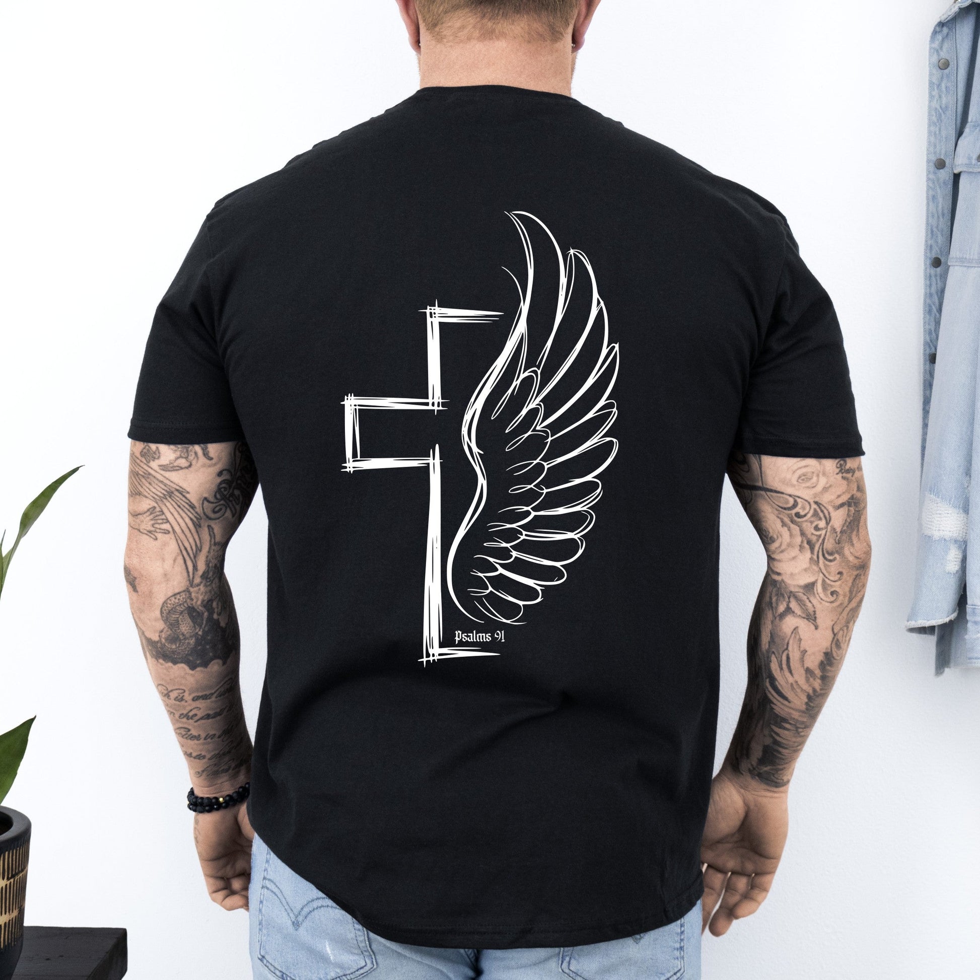 Person wearing a black t-shirt with a winged cross design on the back, standing against a white wall.