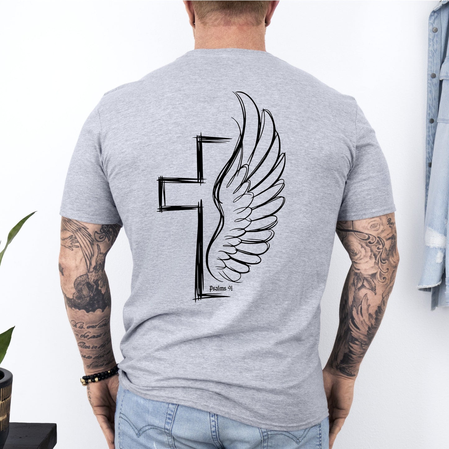 Man wearing a gray t-shirt with a cross and wing design on the back, standing against a white wall.