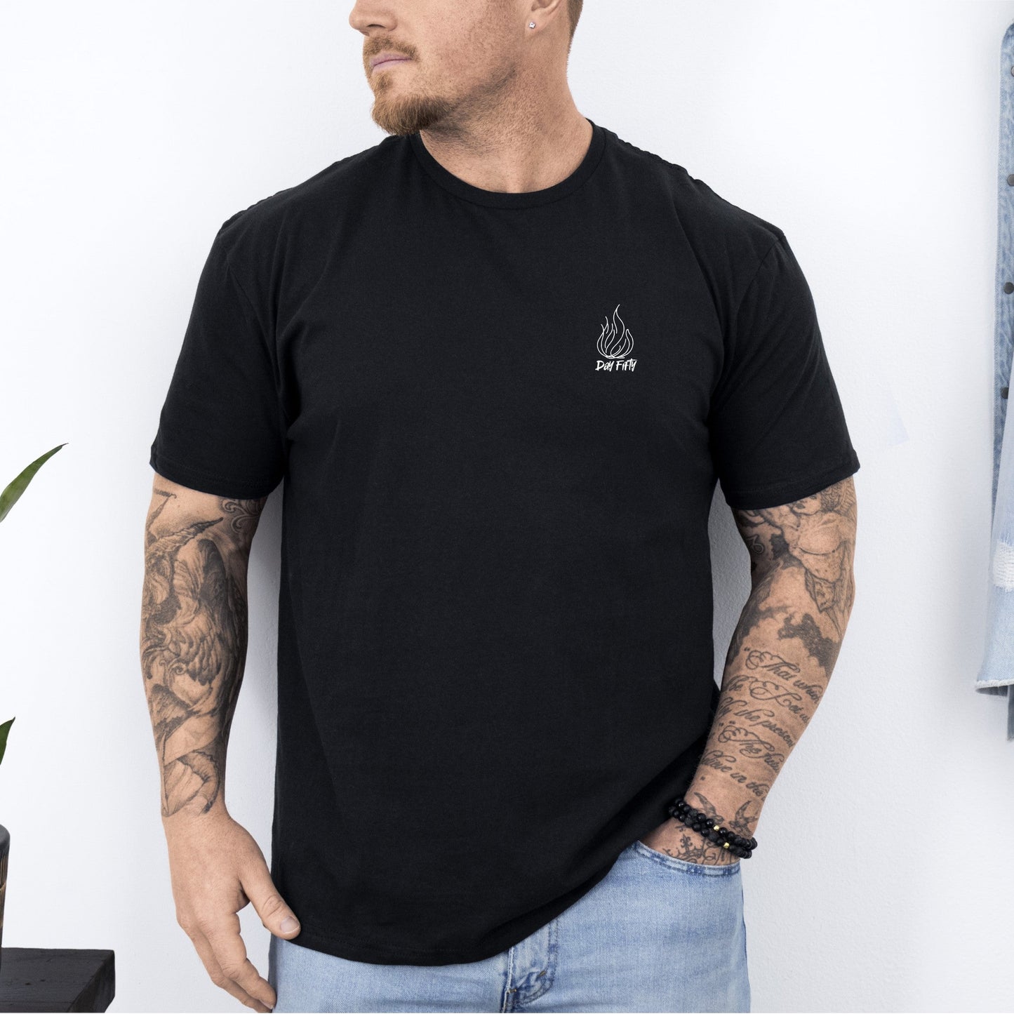 Man wearing a black t-shirt with a logo, standing against a white wall with a plant and jacket in the background.