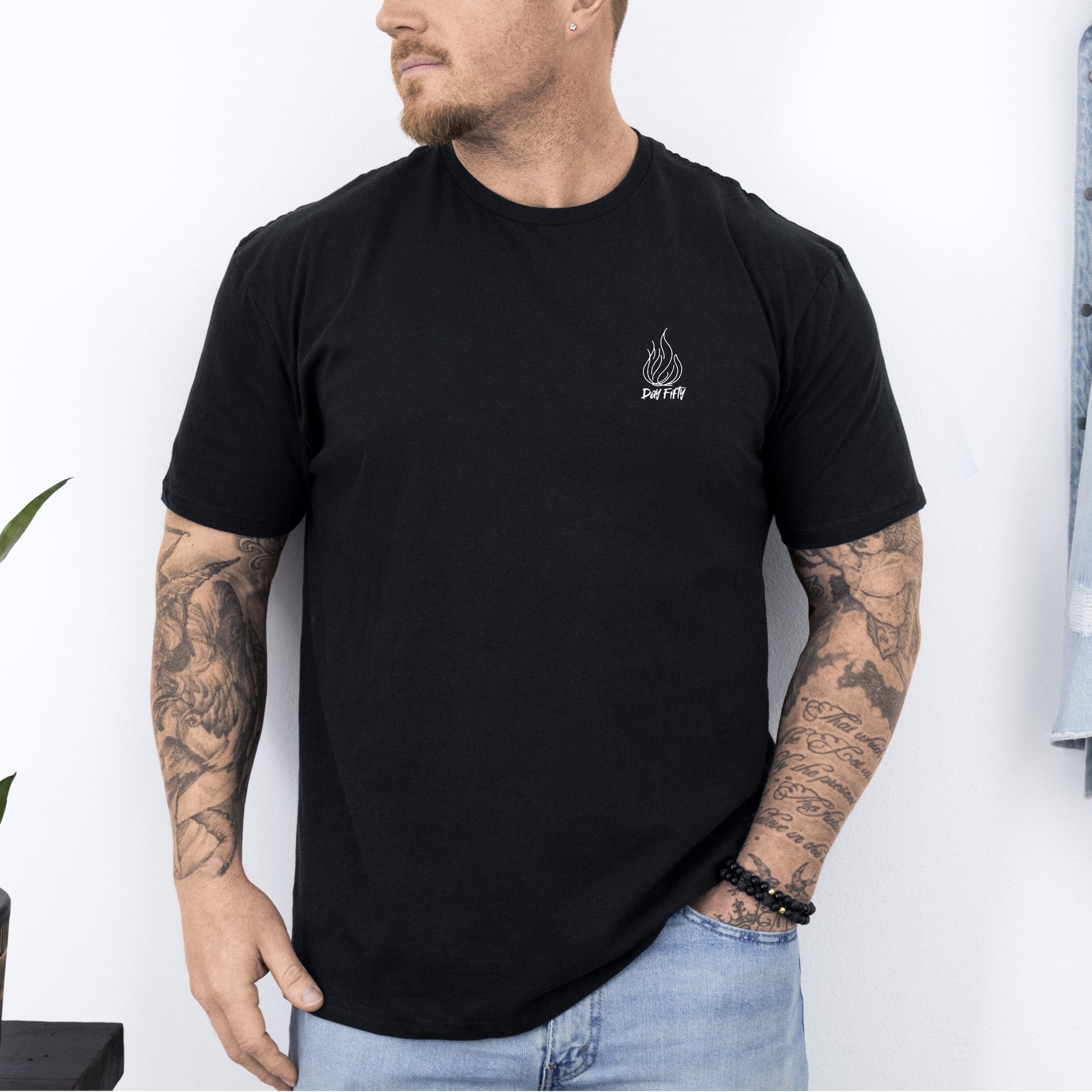 Man wearing a black t-shirt with a logo, standing against a white wall with a plant and jacket in the background.