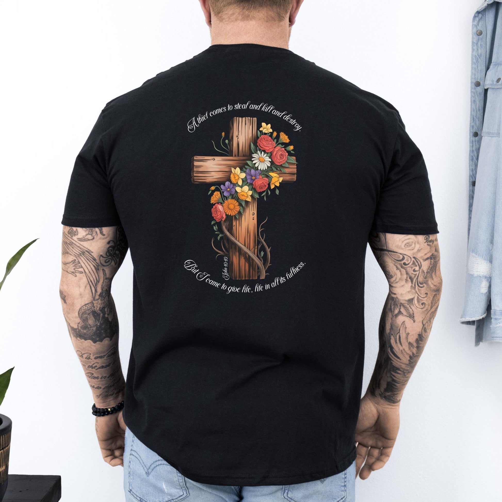 Person wearing a black t-shirt with a wooden cross design on the back, standing against a white wall.