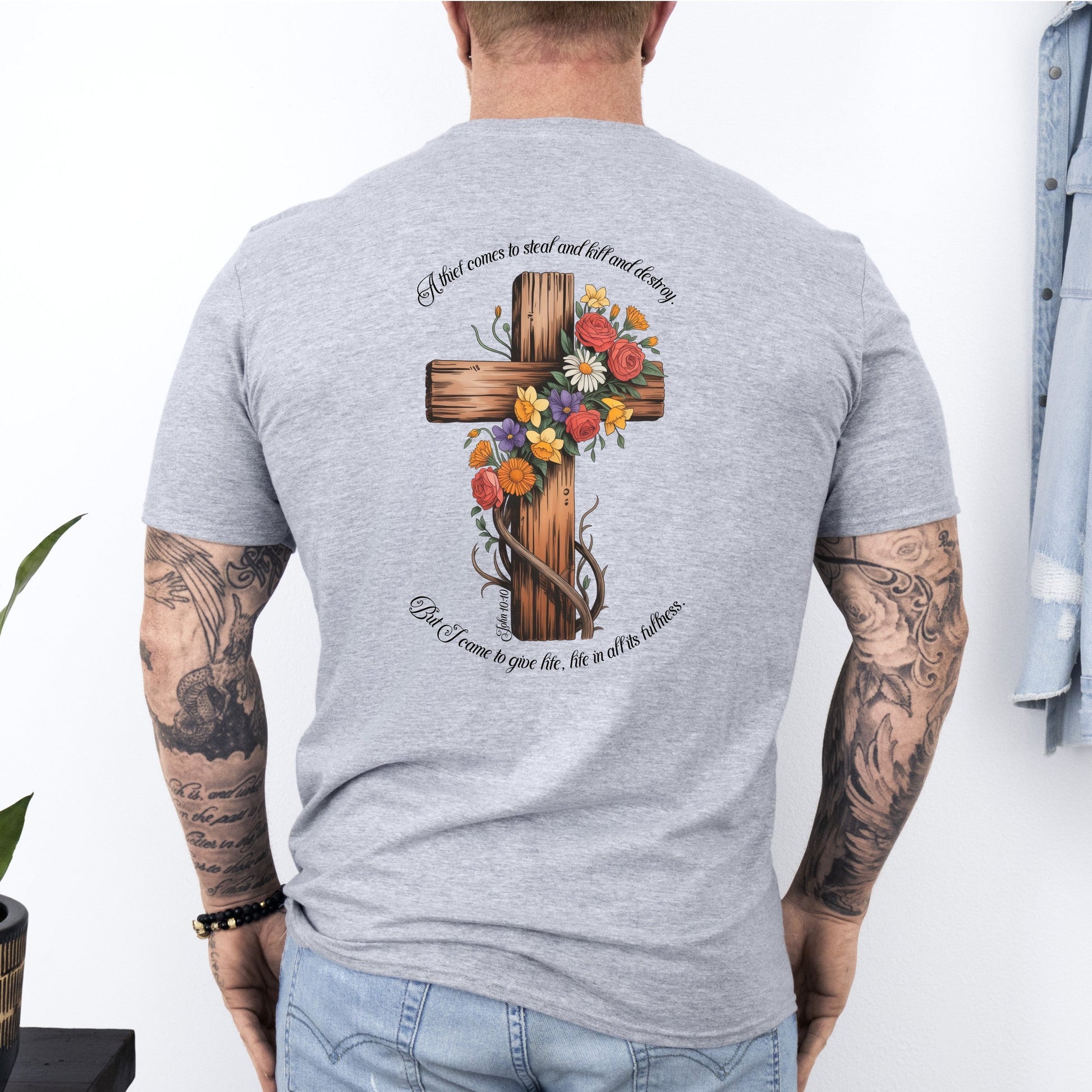 Person wearing a gray t-shirt with a floral cross design on the back, standing against a white wall.